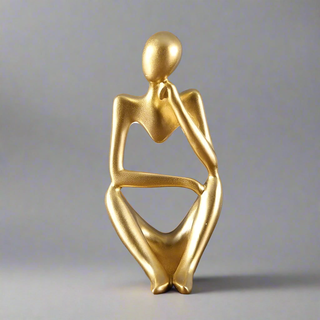 Gold abstract sculpture of a seated figure on a wooden surface with a gray background