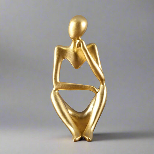 Gold abstract sculpture of a seated figure on a wooden surface with a gray background